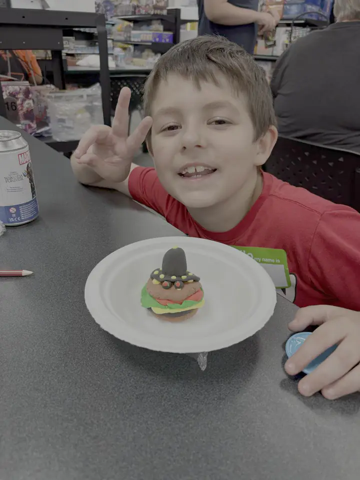 A smiling Kids Club participant flashes a peace sign beside a handmade clay character on a paper plate