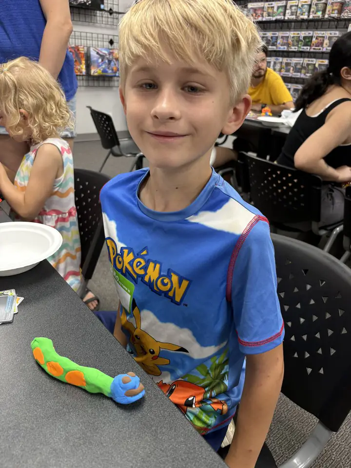 A young Kids Club participant sits at a craft table with a colorful clay creation while other families gather nearby