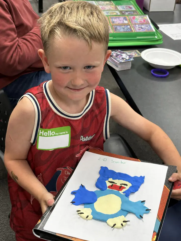 A young Grandma Jane's Kids Club participant holds up a handmade Snorlax craft at a table inside Izzi's Gym
